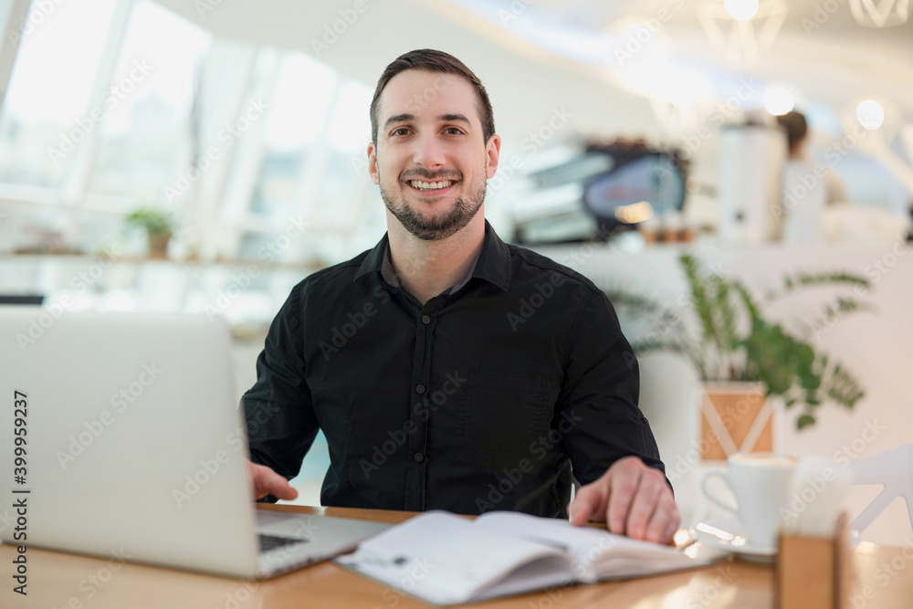Cheerful freelancer man working from cafe. Attractive young man smiling, using his modern laprtop and looking at camera. Cozy coffeehouse on background. Blurred open diary, mug of coffee on table. Cheerful freelancer man working from cafe. Attractive young man smiling, using his modern laprtop and looking at camera. Cozy coffeehouse on background. Blurred open diary, mug of coffee on table.