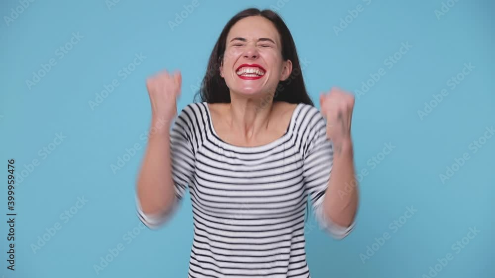 Happy overjoyed brunette young woman 20s years old in striped shirt posing isolated on blue color background studio. People lifestyle concept. Doing winner gesture celebrating clenching fists say yes Happy overjoyed brunette young woman 20s years old in striped shirt posing isolated on blue color background studio. People lifestyle concept. Doing winner gesture celebrating clenching fists say yes