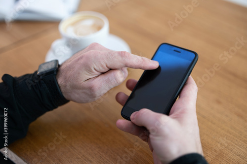 Man using his phone in coffee shop. Smartphone with blank copy space screen. Cup of cappuccino or flat white and open diary on wooden table. Man's hands hodling smartphone.