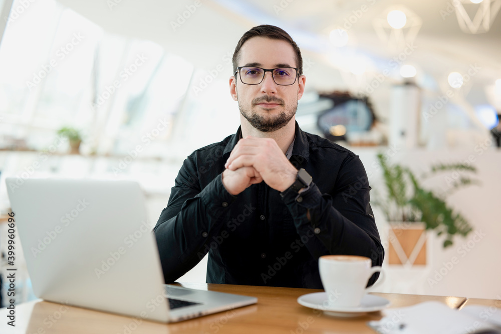 Confident attractive man looking at camera. Modern laptop and cup of cappuccino on wooden table. Light smart coffee shop interior on the background. Freelancer man working in coffeehouse. Confident attractive man looking at camera. Modern laptop and cup of cappuccino on wooden table. Light smart coffee shop interior on the background. Freelancer man working in coffeehouse.