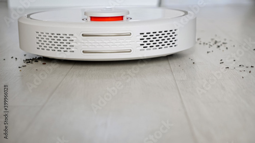 Round automatic robot vacuum cleaner with glowing red lamp drives to trash scattered around wooden floor in brightly lit room closeup