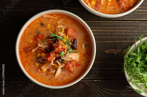 Spicy mexican soup with beans and corn, tomatoes, chicken breast and bean sprouts, bowl with soup and napkin on a dark wooden background, top view