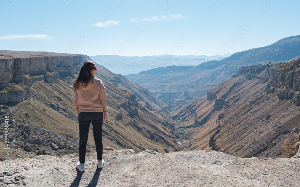 Young brunette woman in loose pink hoodie comes to cliff edge and enjoys picturesque mountain canyon with river on sunny day backside view