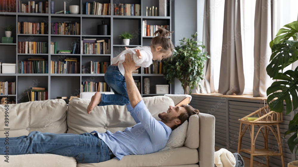 Wide banner panoramic view of smiling young dad rest on sofa at home hold throw in air little daughter. Happy loving father feel playful have fun play with small girl child, relax together on couch. Wide banner panoramic view of smiling young dad rest on sofa at home hold throw in air little daughter. Happy loving father feel playful have fun play with small girl child, relax together on couch.