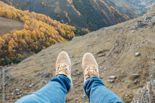 Person in blue pants and beige shoes sits swinging legs on high cliff edge above picturesque valley with yellow trees close upper view