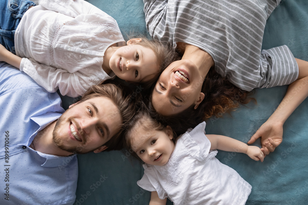 Close up top view portrait of happy parents lying on bed relaxing with cute little daughters. Smiling young Caucasian family with two small girls children have fun rest in bedroom in own home.