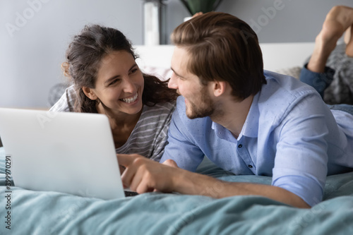 Overjoyed millennial couple lying relaxing in bed shopping online on laptop together. Smiling young Caucasian man and woman rest in cozy bedroom at home browse internet on computer. © fizkes