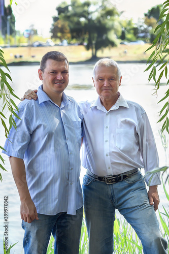 Adult son and elderly father in blue shirts and jeans standing by the lake hugging together. Family relationships. Concept of trust. Two generations of men.