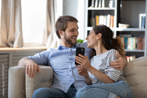 Happy millennial Caucasian couple sit rest on sofa using modern smartphone together. Smiling young man and woman relax on couch in living room at home browsing internet or shopping on cellphone.