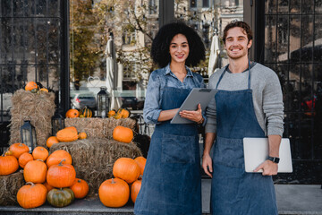 Happy young coffee shop owners standing outdoor near their cafe with laptop and decorating pumpkins preparing for halloween on background.