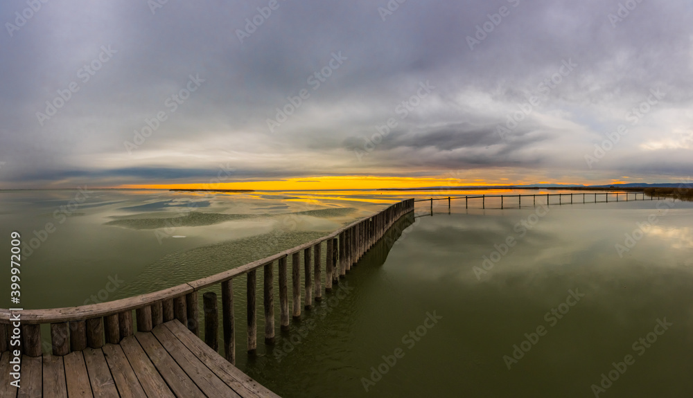long wooden jetty on a frozen lake with sun and rain clouds panorama