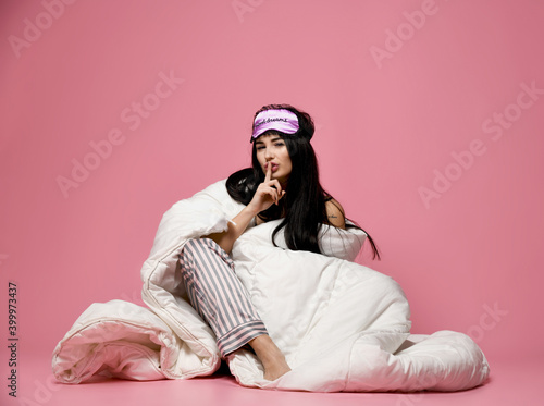 Beautiful brunette girl just woke up with soft blanket and sleepy mask bandage for sleep show tsss shhh quite sign on pink background photo