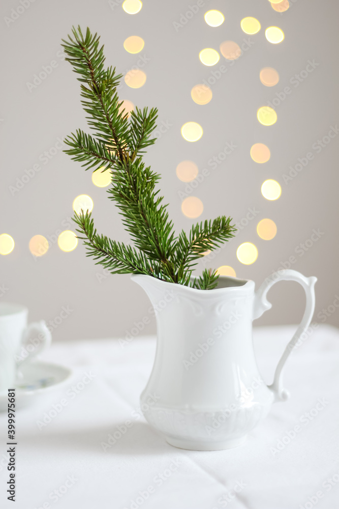 Minimalistic Christmas decoration. A branch of fir tree in a white vintage pitcher and fairy lights in the background Minimalistic Christmas decoration. A branch of fir tree in a white vintage pitcher and fairy lights in the background