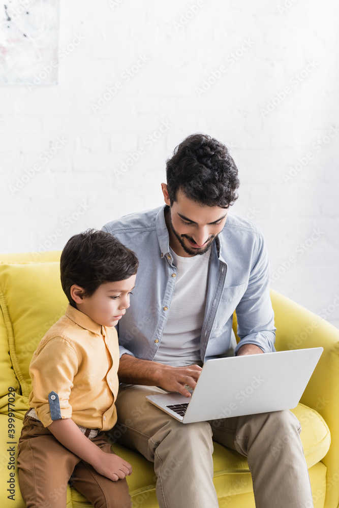 Hispanic father using laptop near son on couch in living room, two generations of men Hispanic father using laptop near son on couch in living room, two generations of men