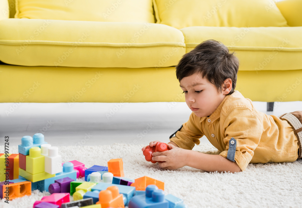 Kid holding building block while lying on floor near couch Kid holding building block while lying on floor near couch