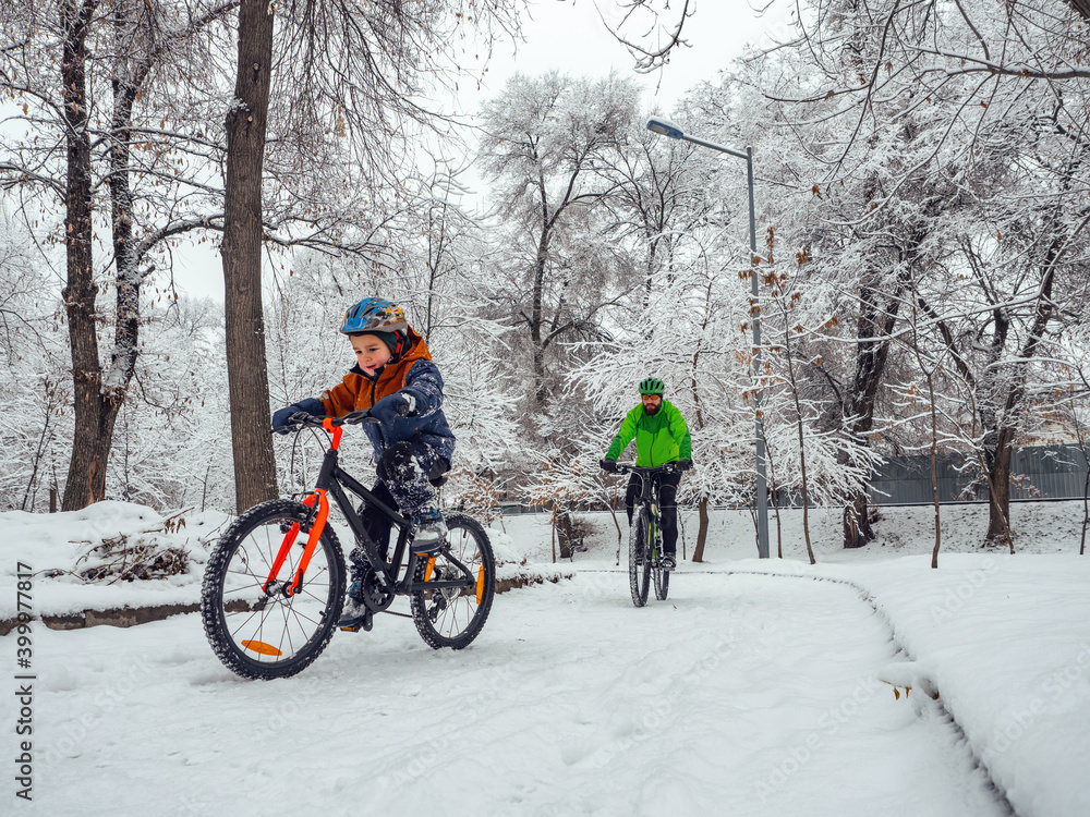 A boy and his father ride bicycles in a winter park. Wonderful winter day outdoors after snowfall. The bearded man is the boy's father, follows him on a bicycle. A boy and his father ride bicycles in a winter park. Wonderful winter day outdoors after snowfall. The bearded man is the boy's father, follows him on a bicycle.