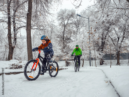 A boy and his father ride bicycles in a winter park. Wonderful winter day outdoors after snowfall. The bearded man is the boy's father, follows him on a bicycle.