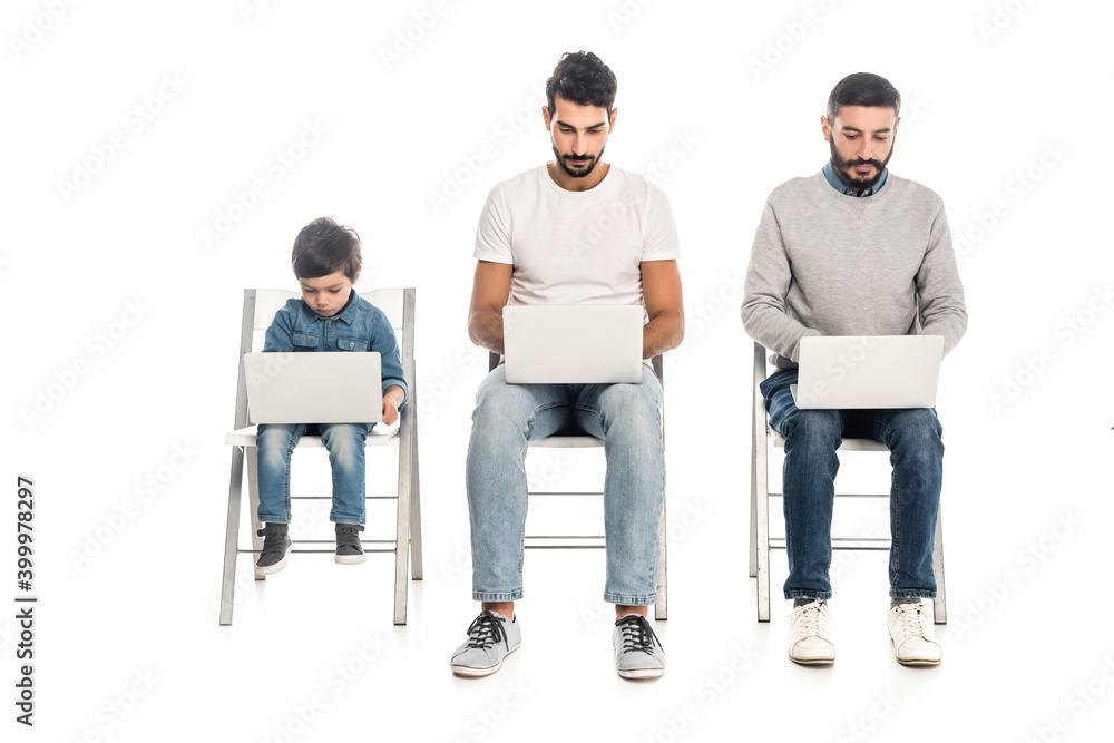 hispanic grandfather, dad and kid using laptops while sitting on chairs on white, three generations of men