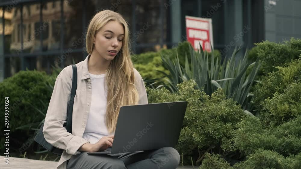 A beautiful young blonde woman is using her laptop sitting outside in the street A beautiful young blonde woman is using her laptop sitting outside in the street