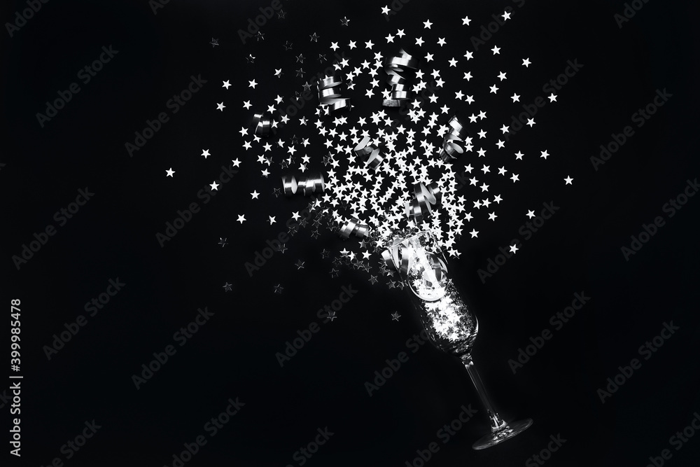 Fototapeta premium Silver star confetti and party streamers poured out champagne glass in flat lay style on black background. Christmas and New Year holiday celebration concept.