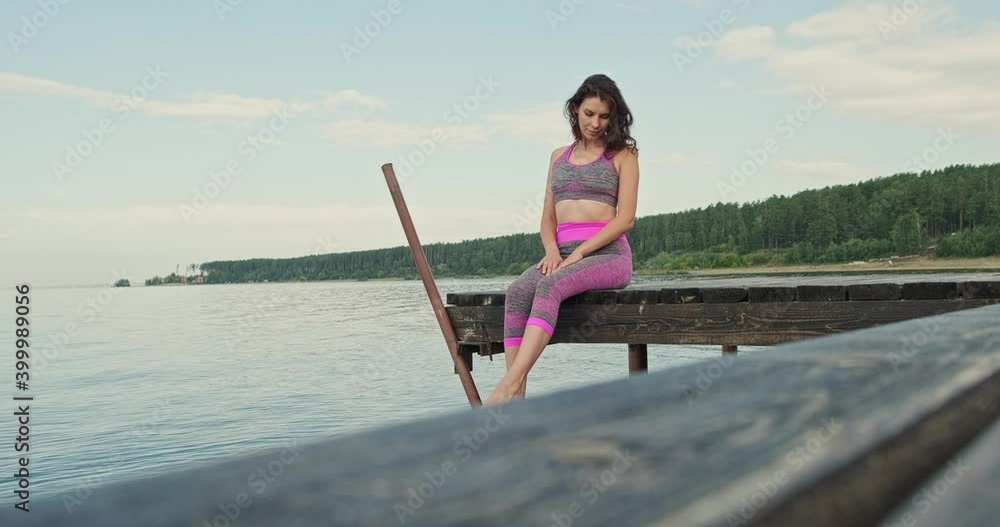 Young girl in tracksuit sits on pier on the seashore, ocean and dangles her legs. Woman sitting on the pier on the berg is resting in the summer by the sea Young girl in tracksuit sits on pier on the seashore, ocean and dangles her legs. Woman sitting on the pier on the berg is resting in the summer by the sea