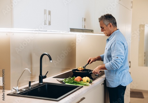 Mature man preparing healthy food for dinner for his family. Concentrated man in casual clothes stands by the stove and stews vegetables in a frying pan. Healthy food concept