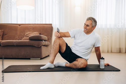 Procrastination during fitness. Senior man resting after home workout with water and phone. Sporty man after practicing yoga, break in doing exercise, relaxing on yoga mat, texting on smartphone.