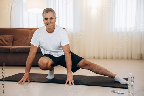 Elderly man doing side lunge exercise. Senior male athlete with fit body warming up, doing leg stretch workout at home. Take care of yourself Modern light room on background. Elderly sport concept.