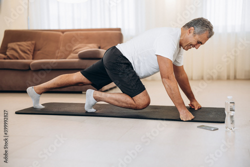 Elderly man doing mountain climber exercises on black yoga mat. Morning workout. Modern living room on background. Plastic bottle of water. Hard workout. Training at home. Elderly sport concept.