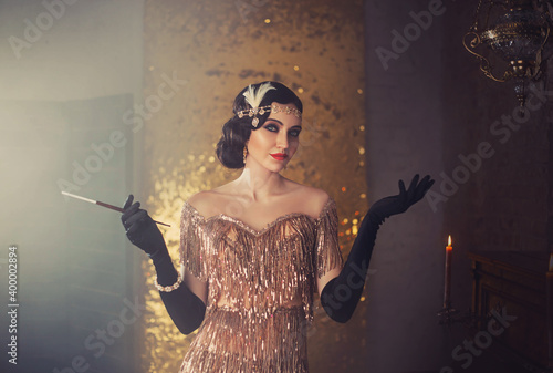 portrait of retro flapper beauty fashion model. Woman holding long slim mouthpiece in hand, cigarette. Party 20s style room full smoke. Gold shiny dress, accessories. Invitation gesture, free space photo