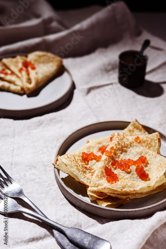 Traditional Russian crepes or pancakes with red caviar on gray plates over greige linen tablecloth. Pancake week or Shrovetide