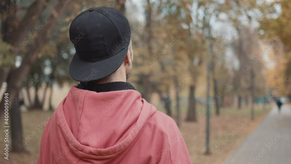 Back shot of man walking in the park, male in black cap wearing red hoodie. Gimbal shot no face of young man on the walk in the park along yellow trees Back shot of man walking in the park, male in black cap wearing red hoodie. Gimbal shot no face of young man on the walk in the park along yellow trees