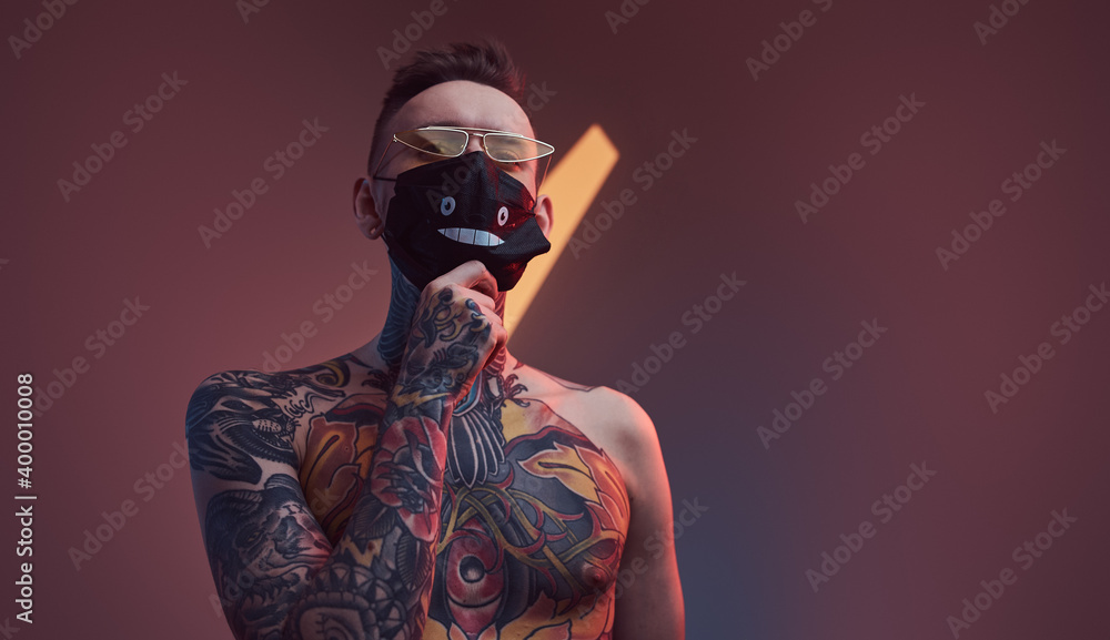 Individual and tattooed macho weared with sunglasses and protective mask poses in dark studio room with spotlight and colourful walls.
