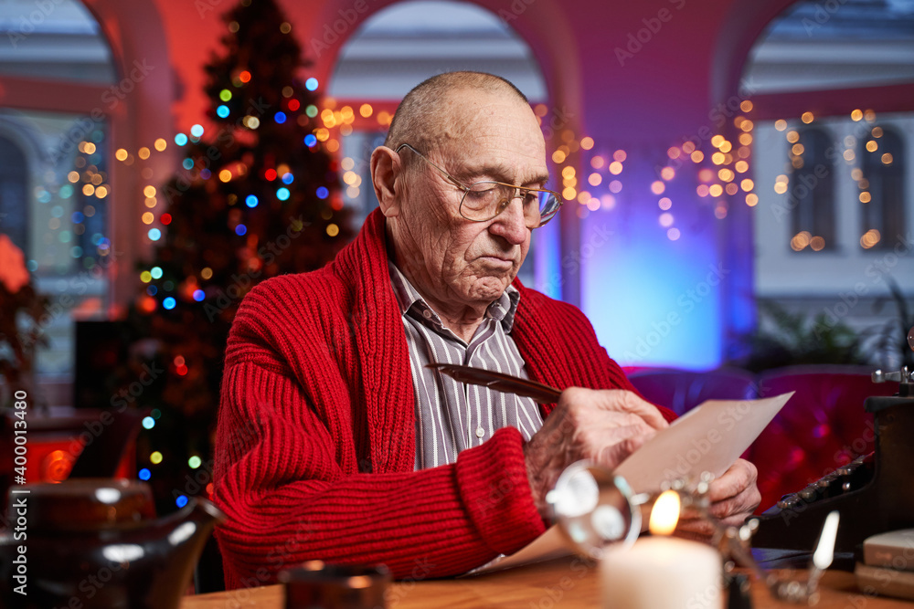 Portrait of a santa with glasses sitting at table and writing christmas cards with serious face