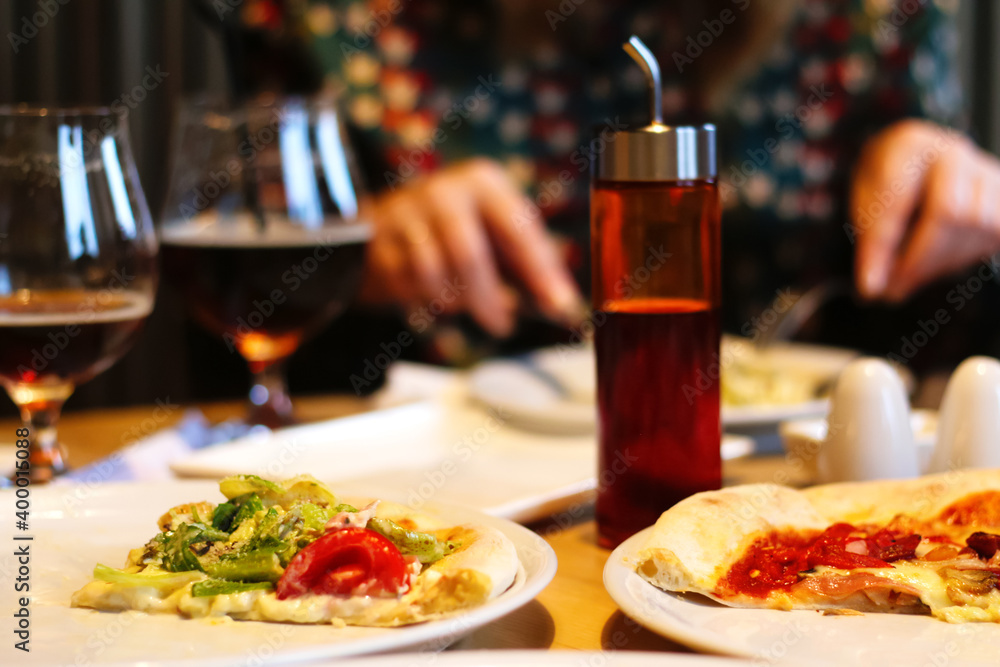 People eat pizza at a table in a cafe.A table in a cafe with pizza. Unfinished pizzas on a two plate. Glasses of beer, olive oil. Blur female hand on background. Selective focus People eat pizza at a table in a cafe.A table in a cafe with pizza. Unfinished pizzas on a two plate. Glasses of beer, olive oil. Blur female hand on background. Selective focus