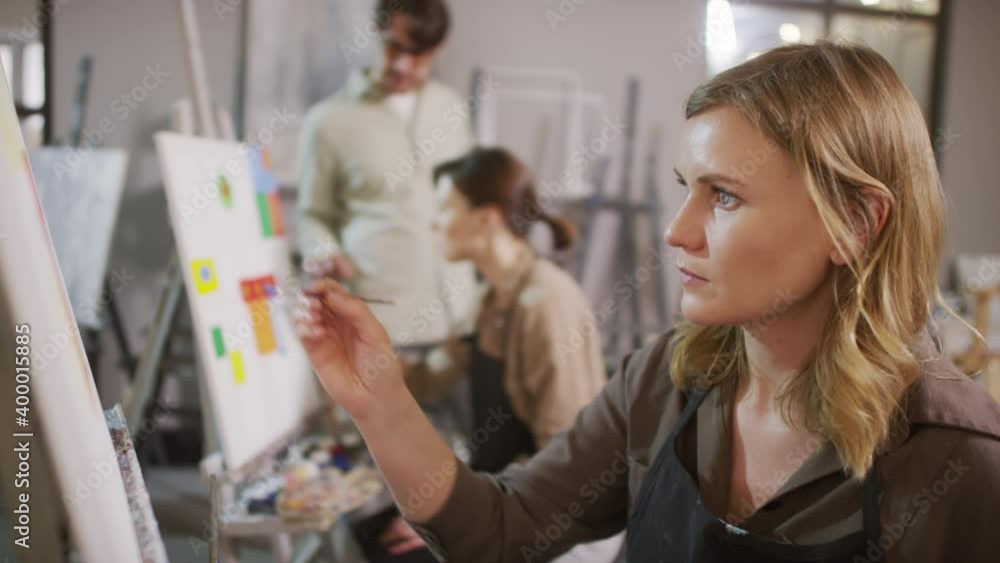 Medium close-up of concentrated pretty woman painting with paintbrush on canvas while male arts tutor talking to student at background in bright modern art studio Medium close-up of concentrated pretty woman painting with paintbrush on canvas while male arts tutor talking to student at background in bright modern art studio