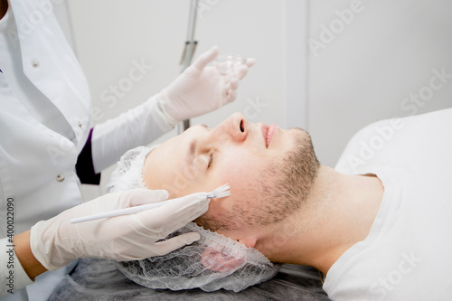 Cosmetologist is putting nutrition lifting on the young man s face in the beauty salon to make it smooth and remove scars and wrinkles