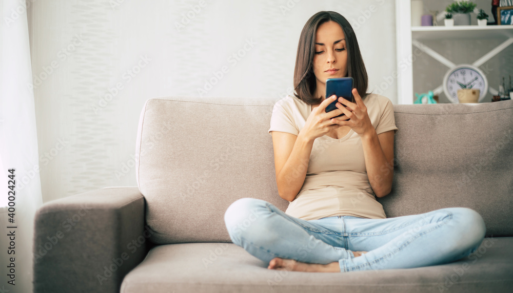 Fototapeta premium Young modern confident smiling woman is working or relaxing at home and sitting on the couch while using her smartphone