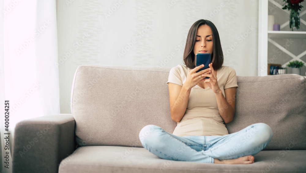 Fototapeta premium Young modern confident smiling woman is working or relaxing at home and sitting on the couch while using her smartphone