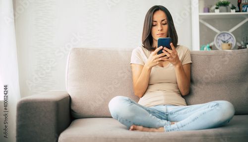 Young modern confident smiling woman is working or relaxing at home and sitting on the couch while using her smartphone