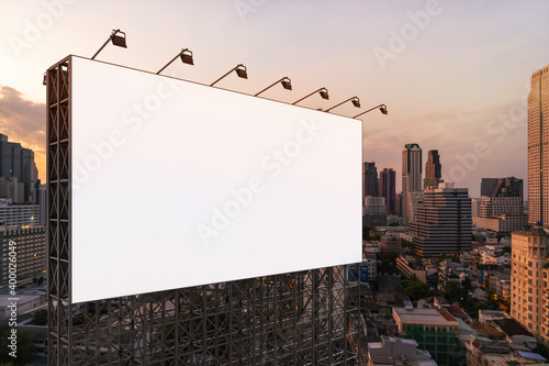 Blank white road billboard with Bangkok cityscape background at sunset. Street advertising poster, mock up, 3D rendering. Side view. The concept of marketing communication to promote or sell idea.