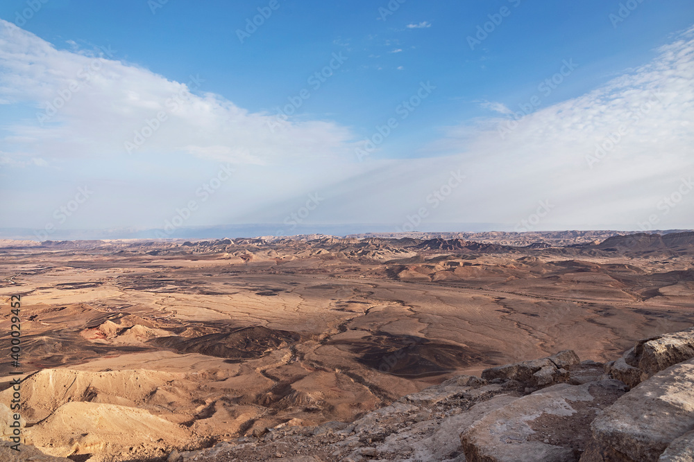 two soft windblown clouds overlap above the maktesh ramon crater in israel with the mountains of jordan in the far background and highly textured rock formations in the foreground