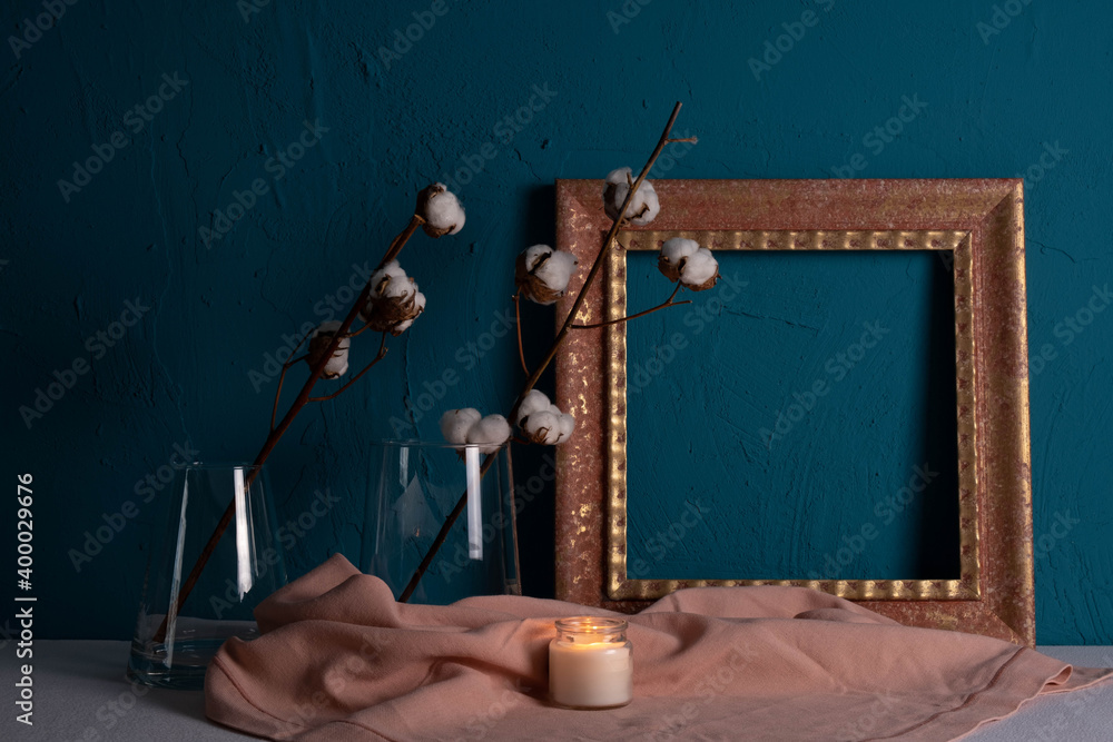 frame for a picture, a candle is burning next and there is a vase with a dry cotton plant on a blue background