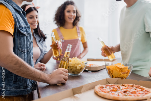 Potato chips and takeaway pizza near hispanic friends with beer at home on blurred background