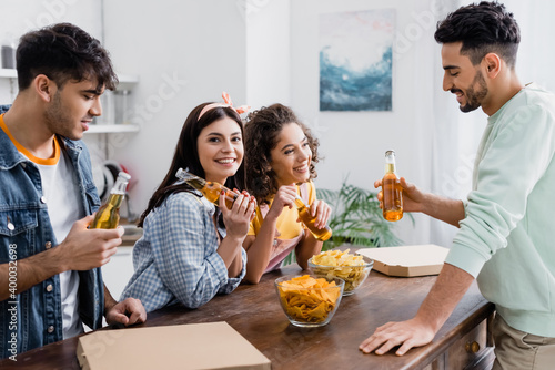 Hispanic woman smiling at camera while holding bottle of beer near friends, pizza and potato chips at home