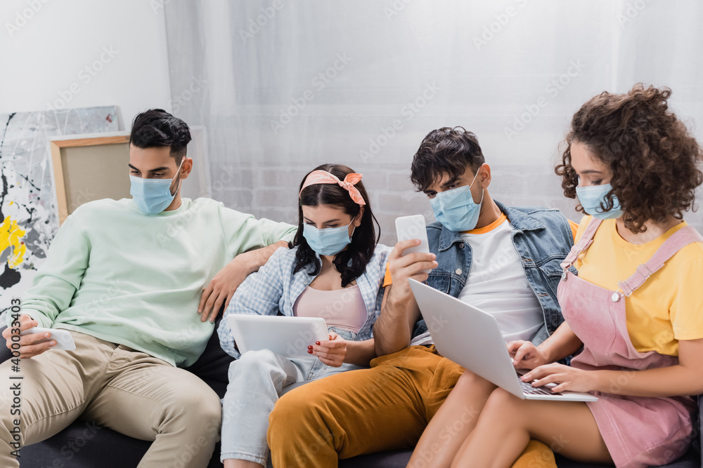 hispanic friends in medical masks sitting on sofa and using gadgets hispanic friends in medical masks sitting on sofa and using gadgets