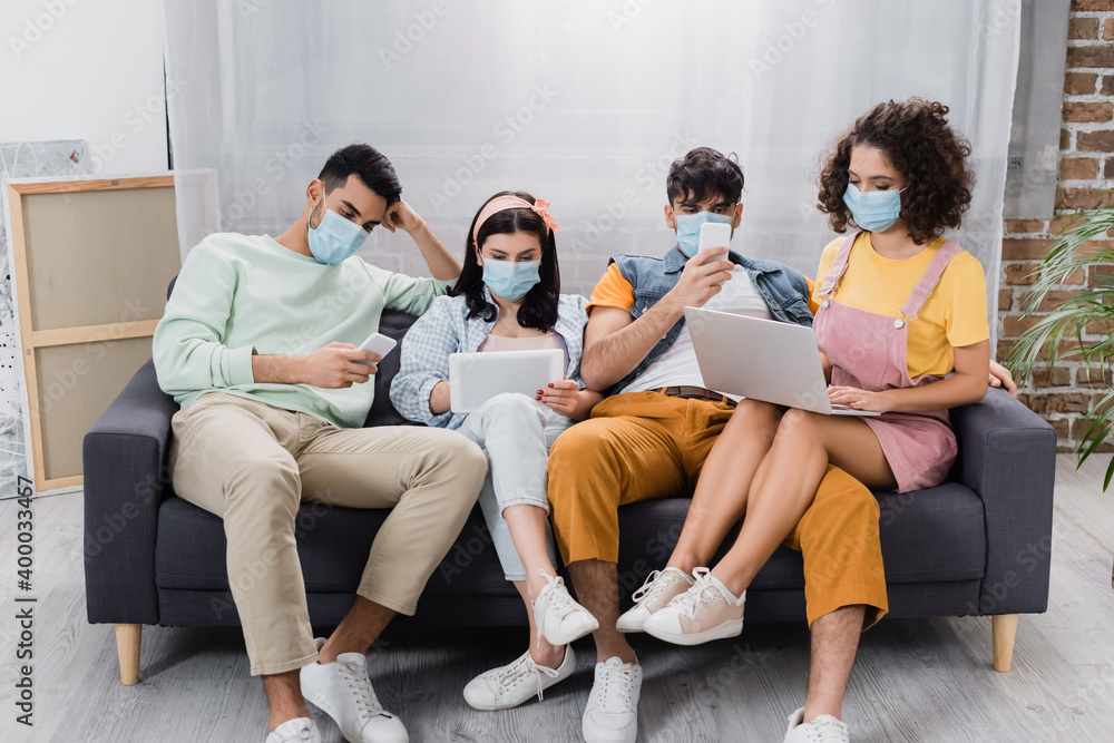 young hispanic friends in medical masks sitting on sofa and using gadgets young hispanic friends in medical masks sitting on sofa and using gadgets