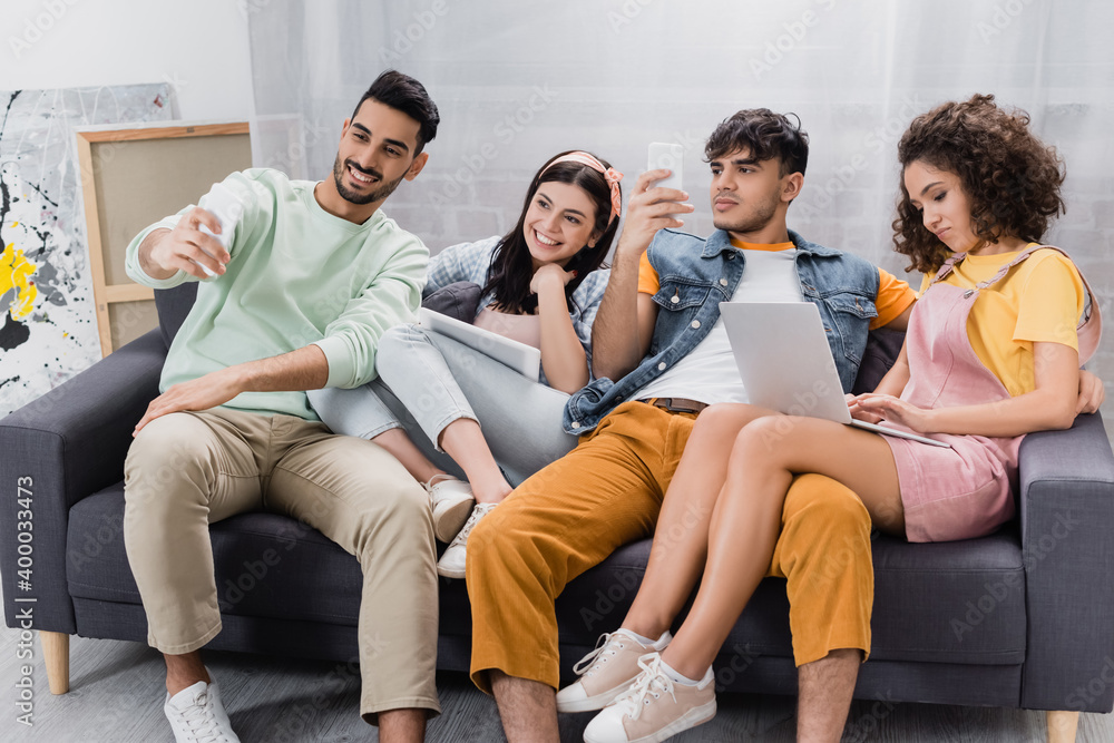cheerful hispanic man taking selfie on mobile phone with friends using gadgets on sofa cheerful hispanic man taking selfie on mobile phone with friends using gadgets on sofa