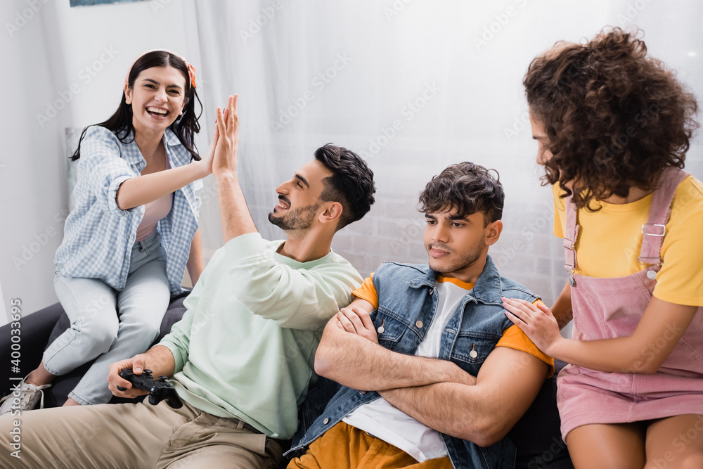 happy hispanic friends giving high five near upset man sitting with crossed arms happy hispanic friends giving high five near upset man sitting with crossed arms