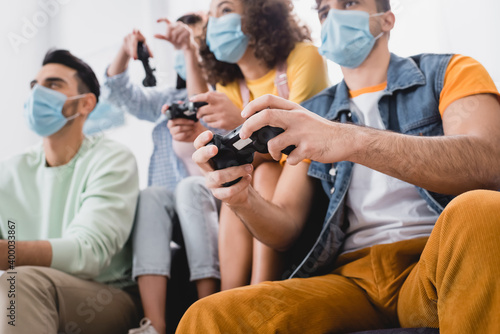 Gamepad in hands of hispanic man in medical mask playing video game with friends on blurred background